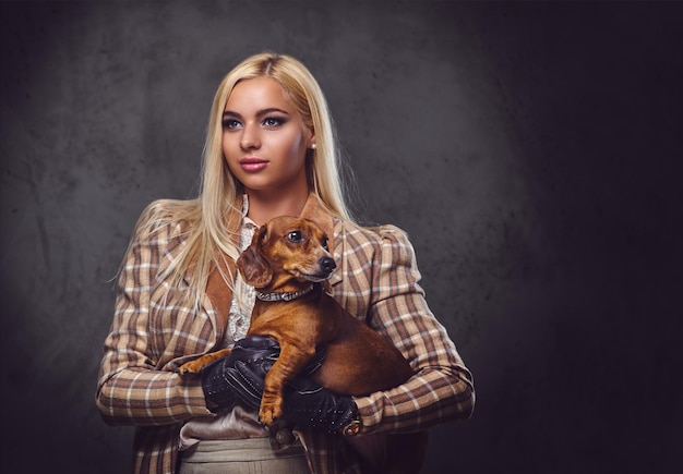 Una elegante mujer rubia vestida con una chaqueta antigua sostiene un perro tejón rojo.