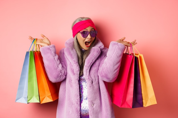 Elegante mujer senior asiática que va de compras, con ropa de moda y gafas de sol, sosteniendo bolsas de la tienda con regalos, date un capricho concepto, fondo rosa.