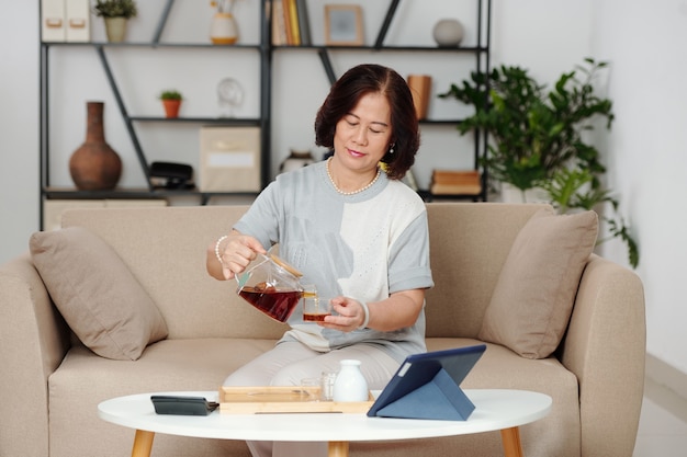 Elegante mujer senior sentada en el sofá en casa y sirviéndose una taza de café