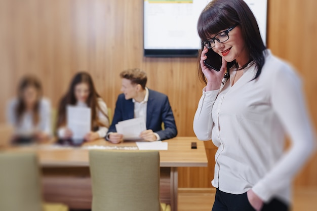 Elegante mujer trabajadora de oficina con gafas con teléfono en manos contra el fondo de colegas de trabajo