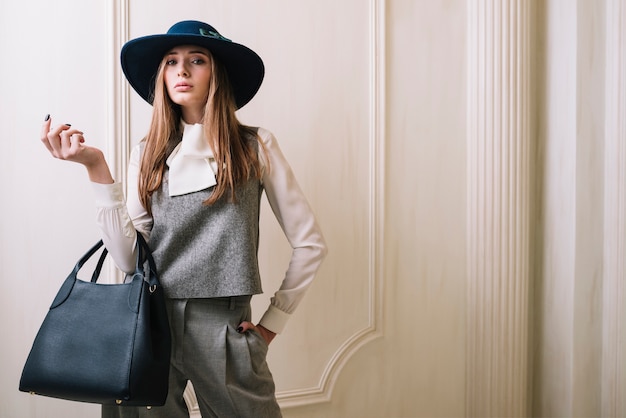 Elegante mujer en traje y sombrero con bolso en habitación.