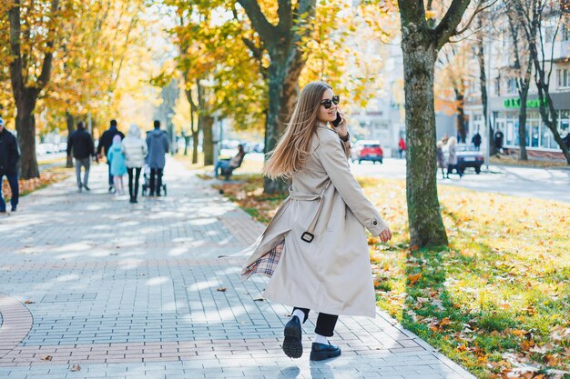 Elegante mulher feliz com casaco de outono e óculos escuros andando ao ar livre e falando no celular mulher alegre falando e conversando no celular ao ar livre enquanto caminhava no parque