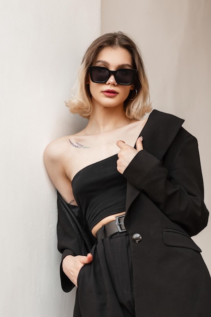 Elegante mulher jovem e bonita com óculos de sol modernos na moda em roupas de negócios pretas da moda com blazer da moda e calças em pé perto da parede branca na rua Estilo urbano feminino