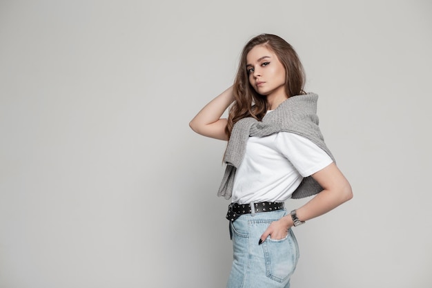 Elegante mulher jovem e bonita com rosto bonito e penteado em roupas casuais da moda com uma camiseta branca jeans azul e um suéter fica no estúdio em um fundo cinza