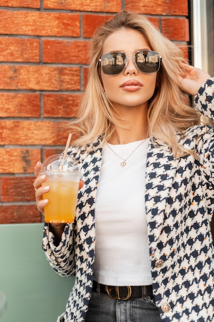 Elegante mulher jovem e bonita em roupas da moda com óculos de sol bebendo uma bebida e descansando na cidade perto de uma parede de tijolo vermelho