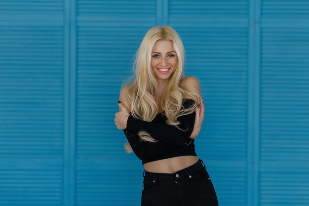 Foto elegante mulher jovem e bonita feliz com um sorriso. cabelo loiro comprido em roupas pretas em uma parede de madeira azul