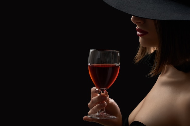 Elegante mysteriöse Frau in einem Hut, der ein Glas Rotwein auf hält