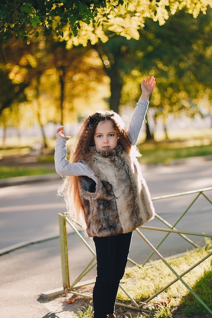 Elegante niña de 4-5 años con botas, abrigo de piel de pie en el parque. Mirando a la cámara. Otoño temporada de otoño