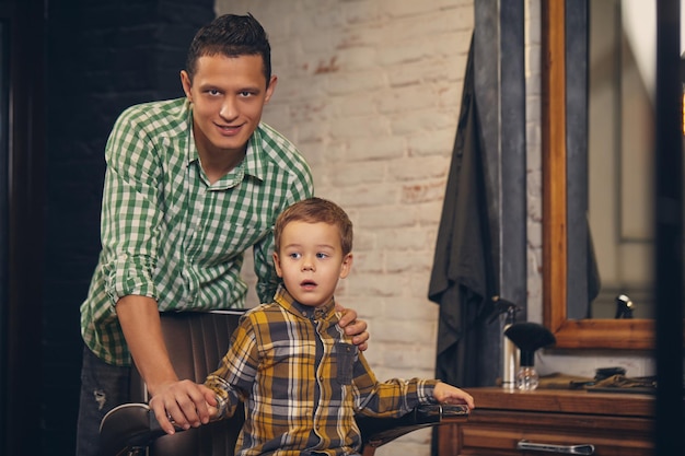 Elegante niño sentado en una silla en la barbería con su joven padre en el fondo, están jugando y divirtiéndose