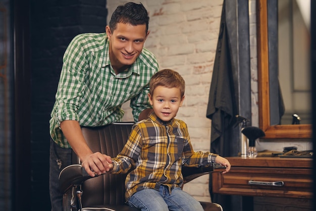 Foto elegante niño sentado en una silla en la barbería con su joven padre en el fondo, están jugando y divirtiéndose