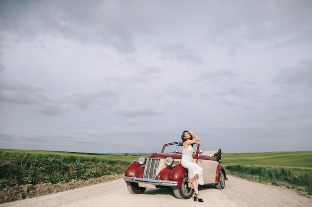 Elegante novia bonita sentada en un auto retro rojo en una carretera de campo