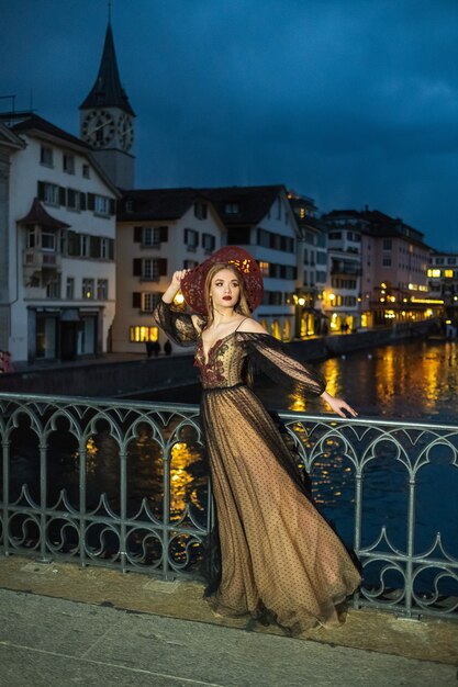 Una elegante novia con un vestido de novia negro y un sombrero rojo posa por la noche en el casco antiguo de Zúrich. Suiza.