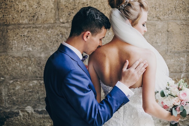 Elegante novio besando suavemente a la hermosa novia con ramo en el hombro en la vista trasera de la calle de la ciudad Hermosa pareja de recién casados abrazándose en edificios antiguos Momento romántico