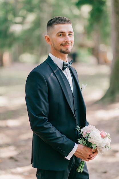 Elegante novio con ramo de flores posando al aire libre.