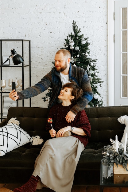 Una elegante pareja de enamorados toma un selfie en su teléfono inteligente con el telón de fondo de un árbol de Navidad en casa