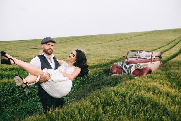 Elegante pareja de novios en un campo verde cerca de coche retro.