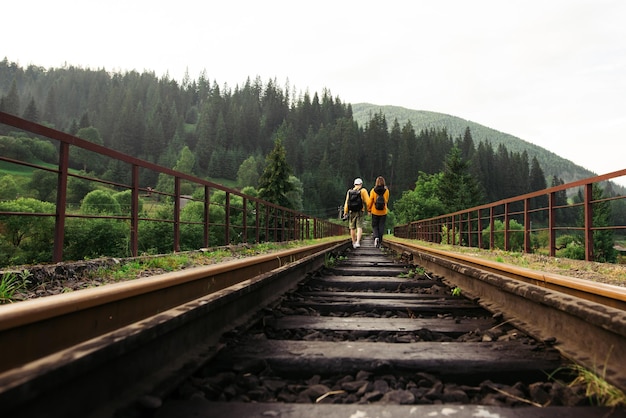 Elegante pareja de turistas con ropa informal camina sobre los rieles en un puente ferroviario en las montañas