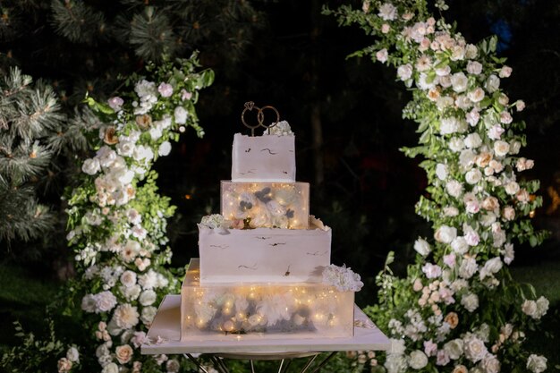 Elegante pastel de boda en niveles iluminado con luces románticas en un telón de fondo floral al aire libre