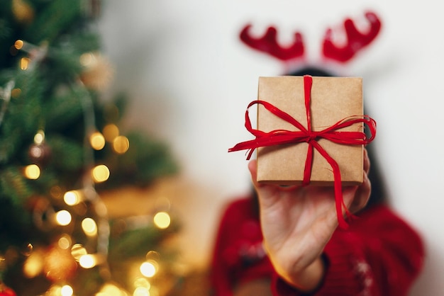 Elegante regalo artesanal con mujer de lazo rojo con sombrero de reno sosteniendo un regalo al frente en el fondo de las luces del árbol de navidad espacio para texto saludos de temporada felices fiestas