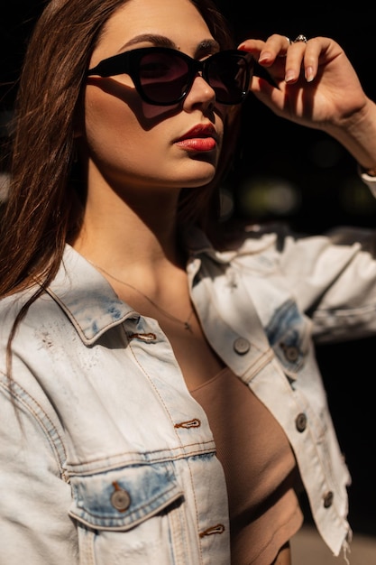 Elegante retrato femenino de una hermosa joven modelo con gafas de sol de moda en una chaqueta de mezclilla informal en la ciudad a la luz del sol