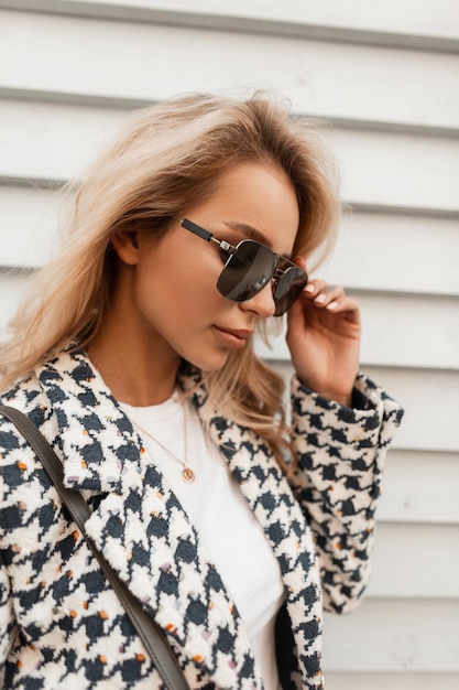 Elegante retrato de mujer bonita rubia con gafas de sol en ropa de moda cerca de una pared vintage de madera