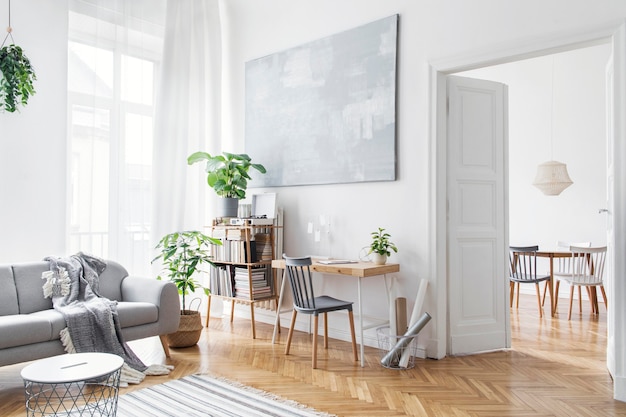 Elegante sala de estar escandinava con muebles de diseño, plantas, atril de bambú, escritorio de madera, cuadros de arte, parquet marrón en la decoración del hogar moderna