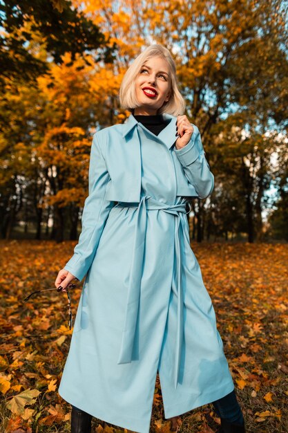 Elegante schöne Geschäftsfrau mit einem Lächeln in einem modischen blauen Mantel mit Sonnenbrille geht in einem Herbstpark mit erstaunlich gefärbtem gelbem Laub spazieren