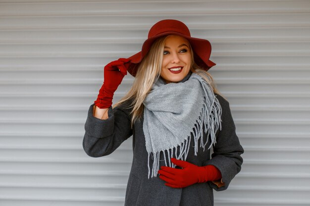 Elegante schöne junge Frau in einem modischen Hut mit einem grauen Schal in einem stilvollen Mantel mit Handschuhen, die nahe einer Holzwand aufwerfen