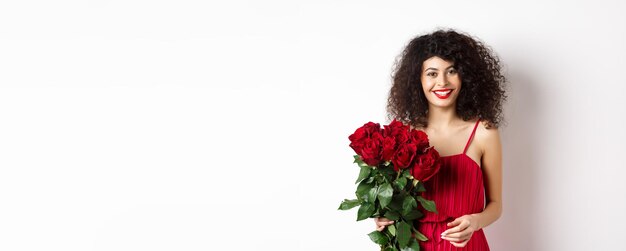 Elegante senhora sorridente com lábios vermelhos e vestido segurando buquê de rosas e olhando feliz backgr branco