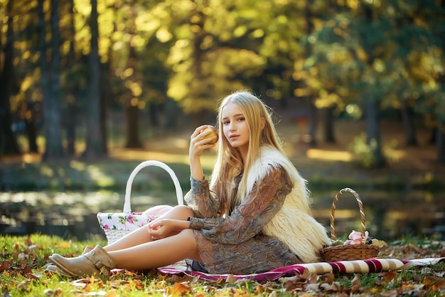 Elegante sensual pensativa linda jovem loira com cabelos longos no vestido de primavera elegante, sentado na manta de piquenique com cesta de frutas no parque ao ar livre na imagem horizontal de fundo natural