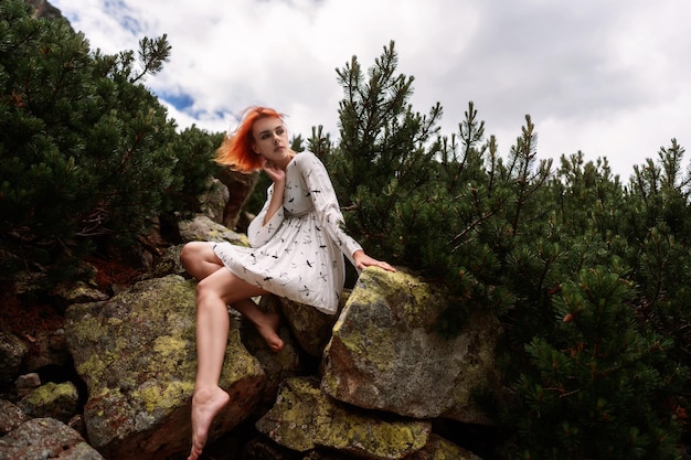 Elegante sexy Dame mit roten Haaren in einem weißen Kleid sitzt auf dem Felsen unter Fichten in den Bergen