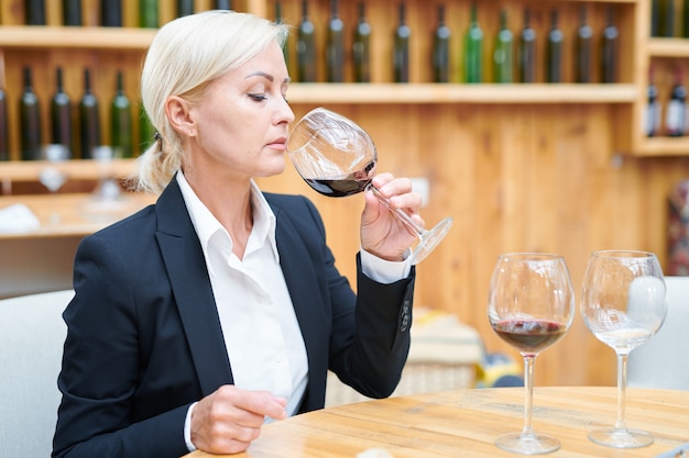 Elegante sommelier sentado à mesa na adega e degustando cabernet em uma taça de vinho para verificar sua qualidade