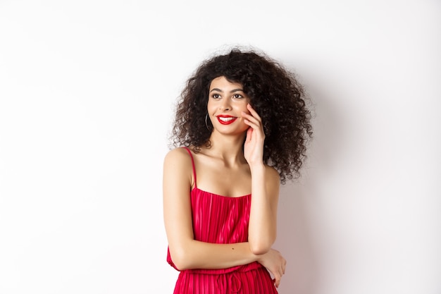 Elegante und romantische Frau in rotem Kleid und Make-up, lächelnd und ihr Gesicht berührend, mit glücklichem Ausdruck beiseite schauend, weißer Hintergrund.