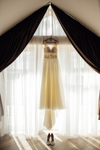 Un elegante vestido de novia cuelga junto a una ventana con cortinas Cerca hay zapatos de mujer