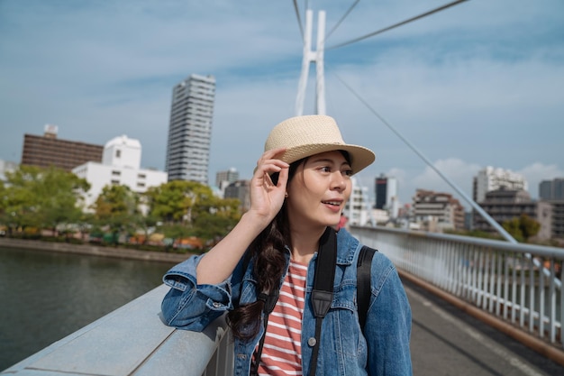 elegante viajera parada en el puente en un día soleado mirando a un lado. joven turista con sombrero de paja visitando la ciudad de osaka. edificios altos en la ciudad sobre fondo de sol.