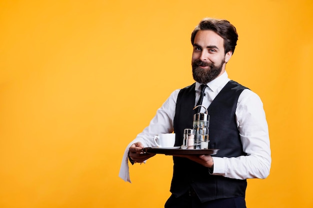 Eleganter Butler hält ein Tablett in der Hand, um Salz und Pfeffer auf den Tisch im Restaurant zu legen, und trägt eine Tasse Kaffee auf einem Teller für die Kunden im Studio. Professioneller luxuriöser Kellner posiert vor der Kamera.