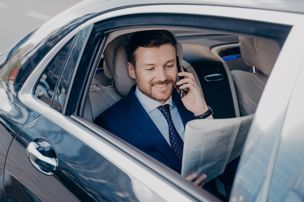 Eleganter Geschäftsmann in formeller, stilvoller Kleidung, der in einem Luxusauto reist, Zeitung liest und Finanzinformationen sucht, während er mit dem Handy spricht, zufrieden lächelt, um gute Nachrichten zu hören