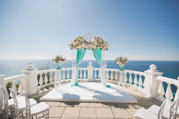 Eleganter Hochzeitsbogen mit frischen Blumen, Vasen auf Ozean und blauem Himmel.