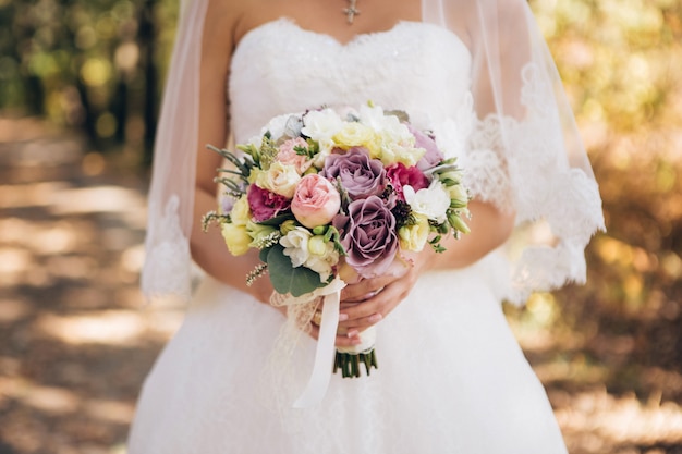 Eleganter Hochzeitsstrauß in den Händen der Braut. Blumenstrauß mit lila, weißen, rosa Rosen. Hochzeitsstrauß im Herbstwald