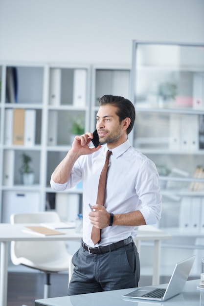 Eleganter junger Agent, der Kunden per Smartphone berät, während er im Büro am Schreibtisch steht