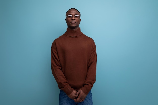 Eleganter junger amerikanischer Brunet-Mann in stilvollem braunem Pullover posiert vor isoliertem blauem Hintergrund mit