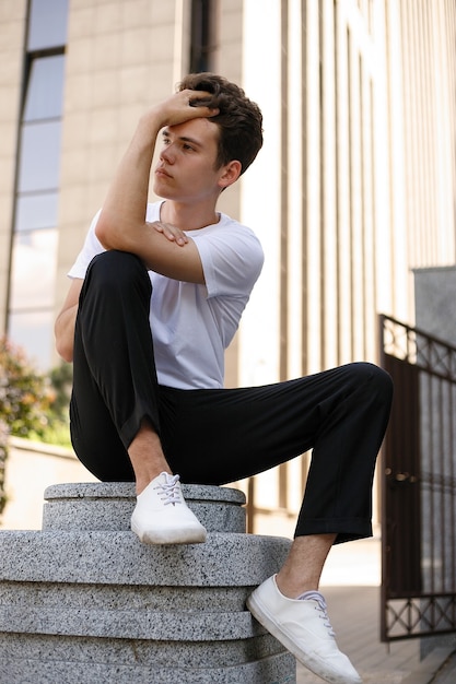 Eleganter junger Mann in einem modischen schwarzen Hemd in einem weißen, stylischen T-Shirt in schwarzer Hose mit trendiger Frisur ruht in der Nähe eines modernen Geschäftszentrums. Attraktiver Kerl auf der Straße an einem Sommertag.