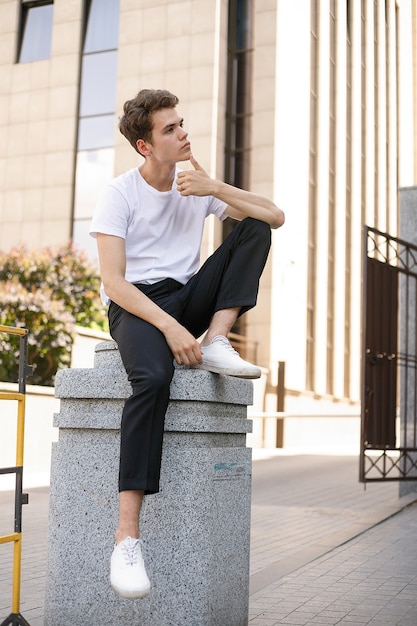 Eleganter junger Mann in einem modischen schwarzen Hemd in einem weißen, stylischen T-Shirt in schwarzer Hose mit trendiger Frisur ruht in der Nähe eines modernen Geschäftszentrums. Attraktiver Kerl auf der Straße an einem Sommertag.