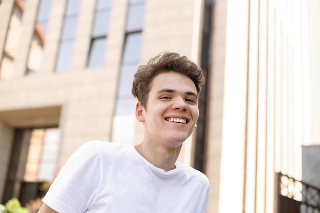 Eleganter junger Mann lächelt in einem modischen schwarzen Hemd in einem weißen stylischen T-Shirt in schwarzer Hose mit trendiger Frisur liegt in der Nähe eines modernen Geschäftszentrums. Nahaufnahmeportrait