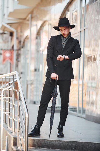 Eleganter junger Mann mit schwarzem Hut und Regenschirm im Freien in der Stadt