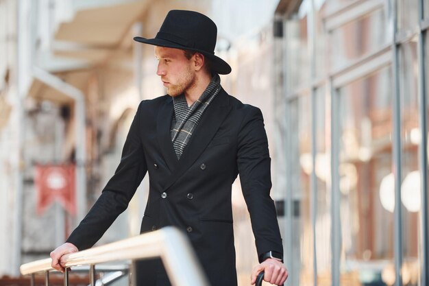 Eleganter junger Mann mit schwarzem Hut und Regenschirm im Freien in der Stadt