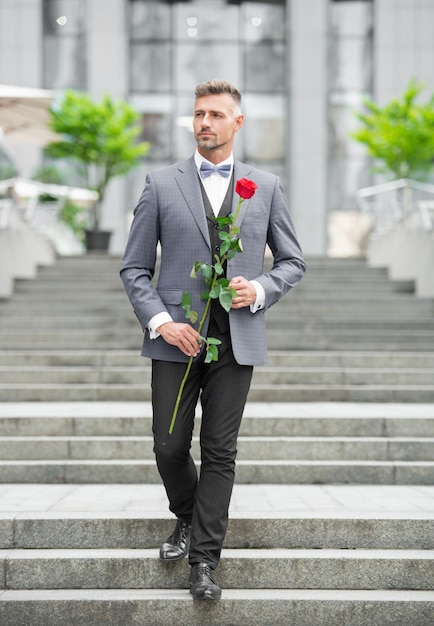 Eleganter Mann im Smoking. Mann mit Smoking-Fliege. Spaziergang im Freien. Hübscher Smoking-Mann mit roter Rose