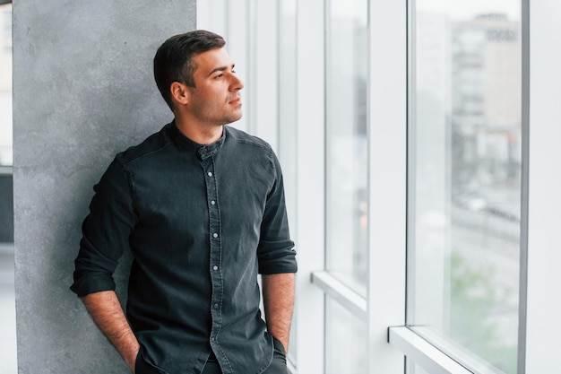 Eleganter Mann in schwarzem Hemd, der sich drinnen an die Wand lehnt und durch das große Fenster schaut