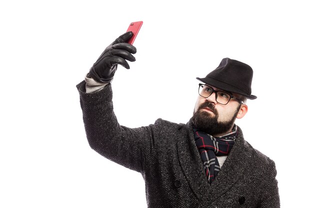 Eleganter Mann mit Bart, gekleidet in einen eleganten Mantel und Hut macht Selfie. Über weißer Wand isoliert.