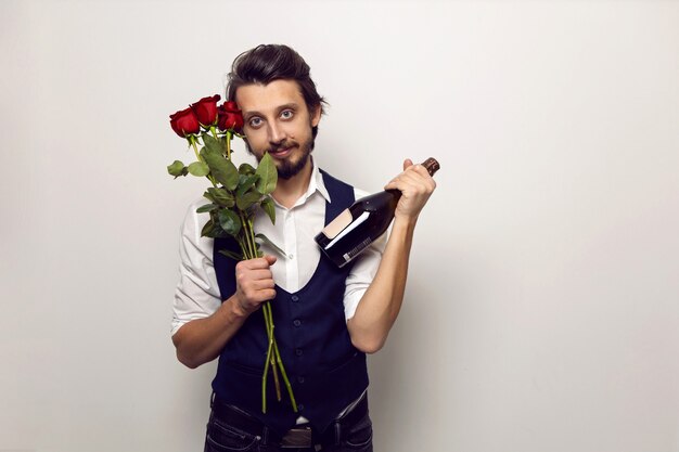 eleganter Mann mit Bart und Brille am Valentinstag in einem weißen Hemd und Weste an einer weißen Wand steht mit einem Strauß roter Rosen und einer Flasche Champagner